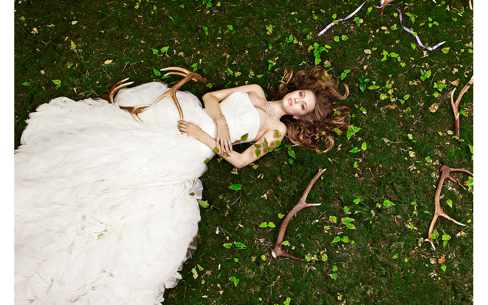 Model laying down in green moss in a white dress holding antlers wearing jewellery in a jewellery advertising photo shoot for winsor bishiop shot in Narford Norfolk