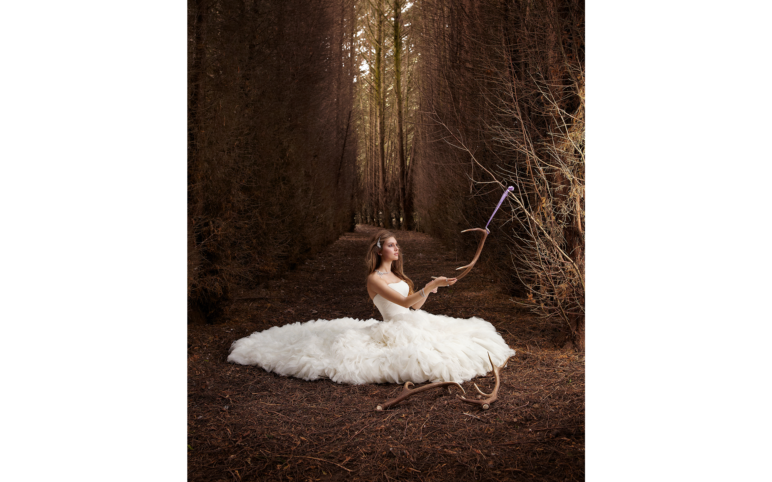 Model sitting down in a forest holding deer antlers looking at bright light coming from the trees