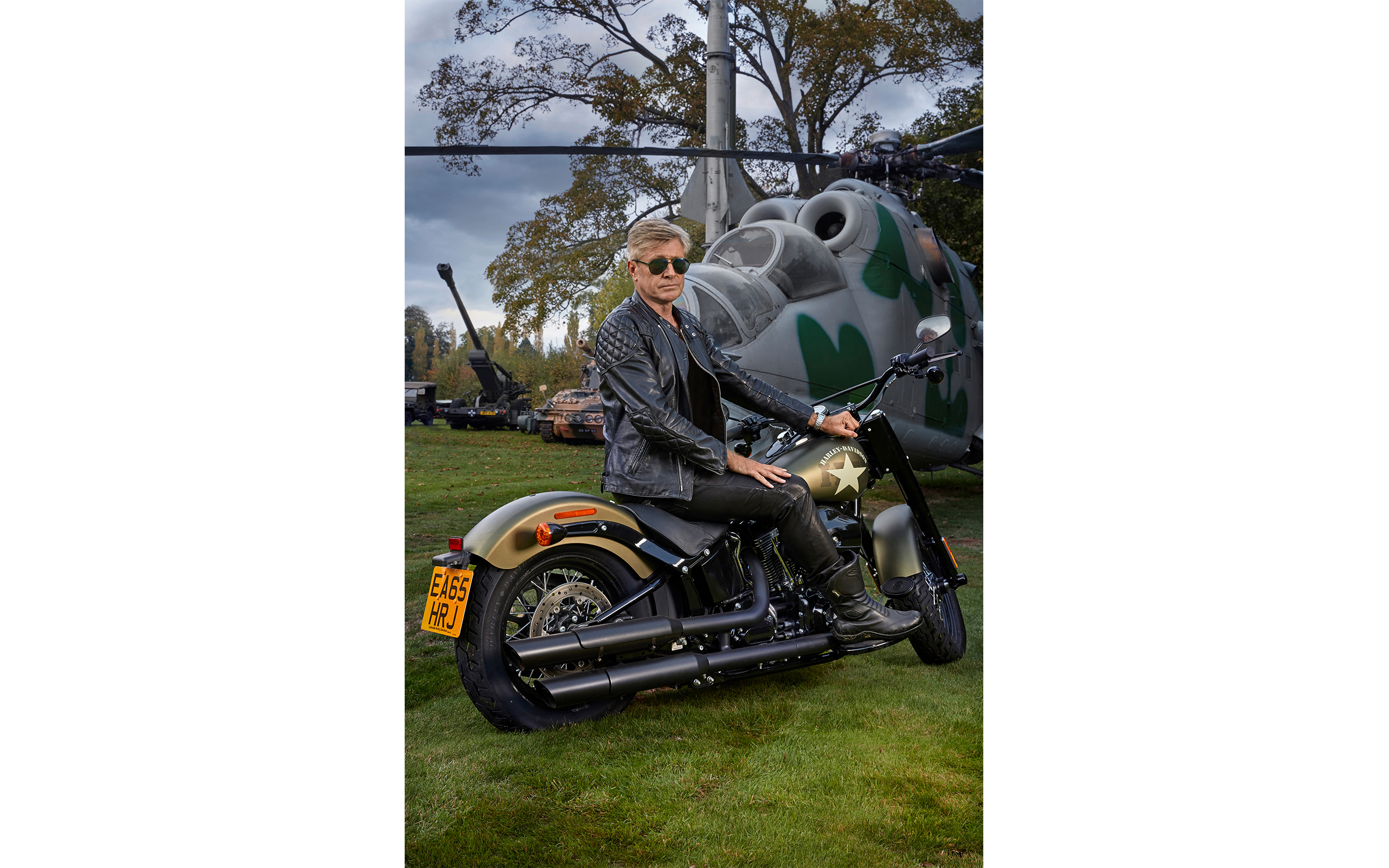 Photo shoot portrait of Man sitting on a Harley Davison motorbike front of a helicopter and tanks in a large garden in London